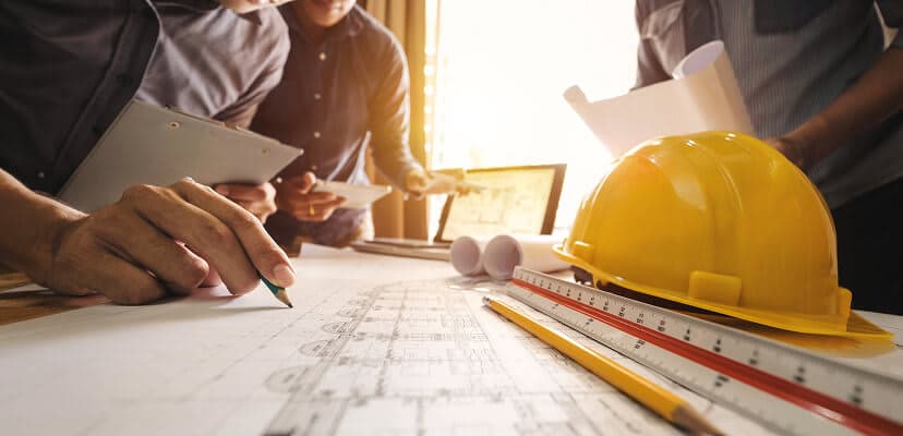 A worker looking over construction plans spread out over a desk with a ruler and hard hat next to it
